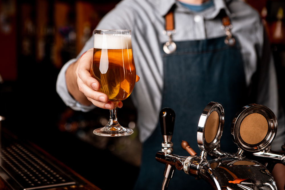 Beer friday evening. Barman gives glass of light beer with foam in dark interior